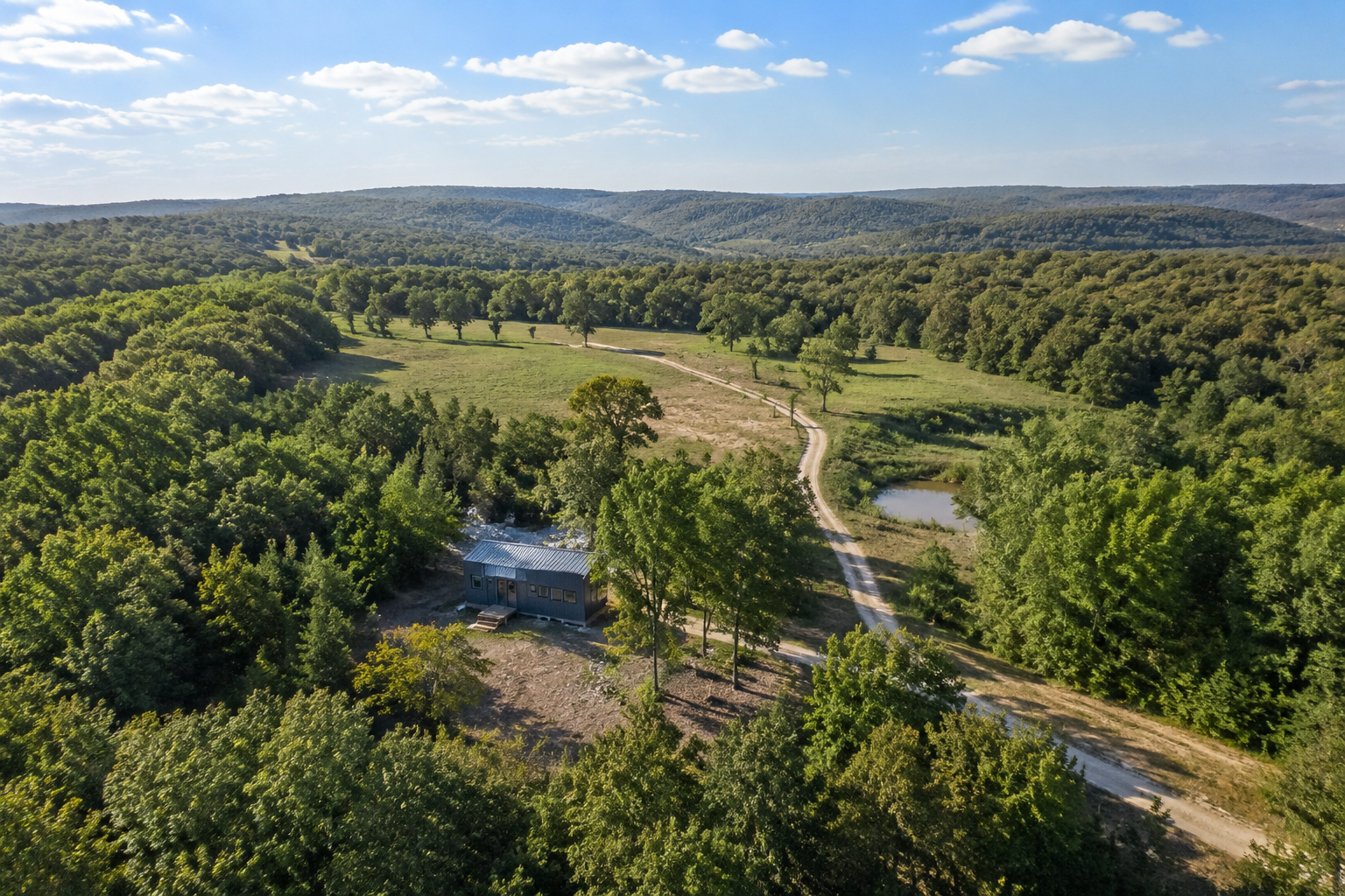 Aerial drone view of affordable rural Arkansas land with a tiny home on a large wooded lot, Ozark hills in background, blue
